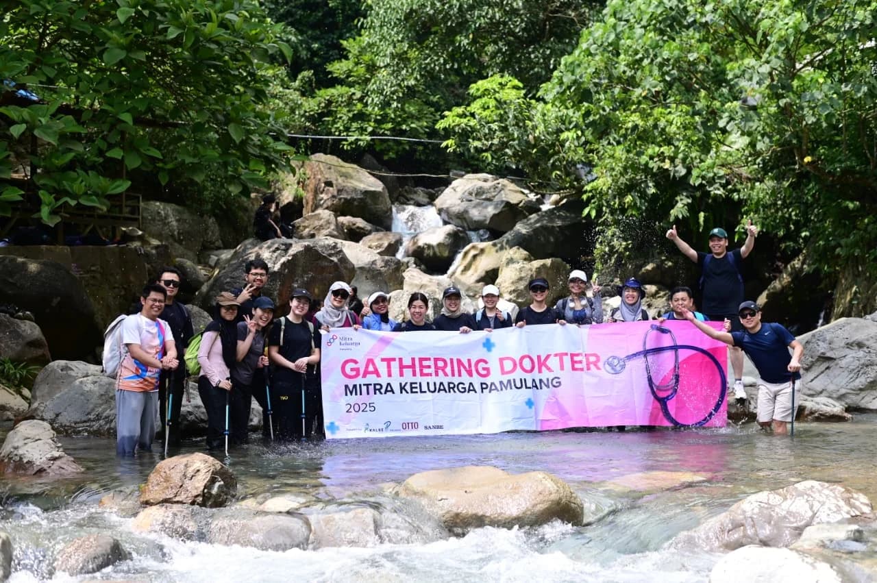 Seorang peserta wanita sedang trekking menggunakan tongkat di jalur yang dikelilingi rerumputan tinggi.