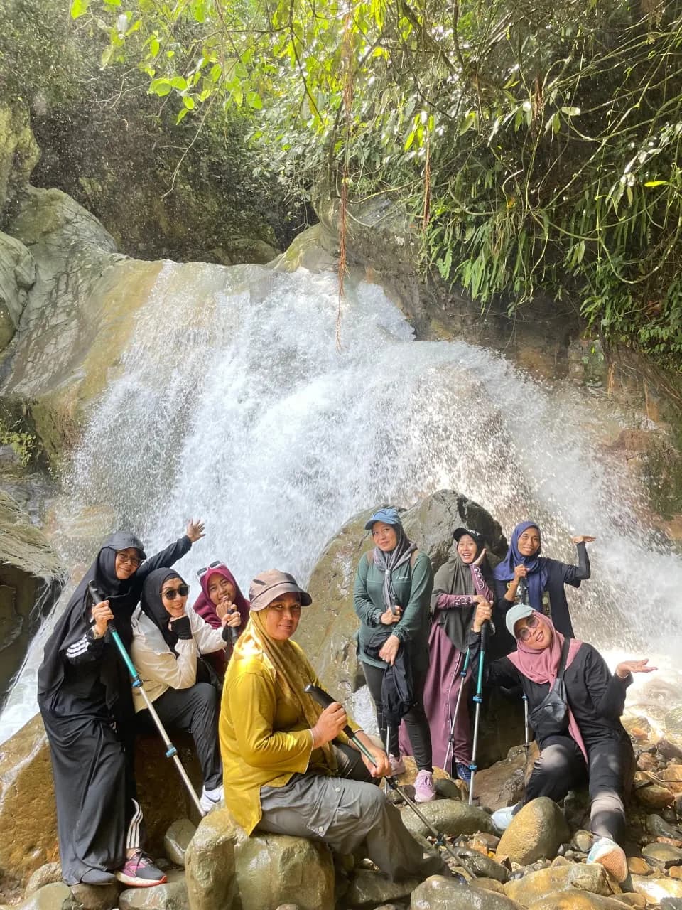 Sekelompok peserta wanita berpose ceria di bebatuan depan air terjun yang deras.